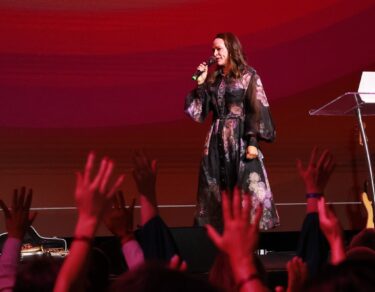 Bispa Raquel Moreira pregando no palco do Modeladas Mais 2026 com as mãos do público erguidas