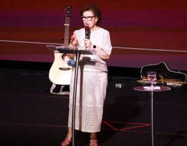 Dra. Ilma Cunha pregando no palco do Modeladas Mais 2026, com o violão e o telão ao fundo