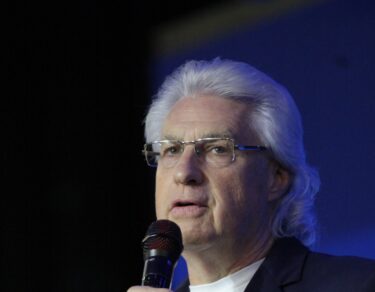 Charles Scott, de cabelos brancos e óculos, fala ao microfone na Conferência Global 2026