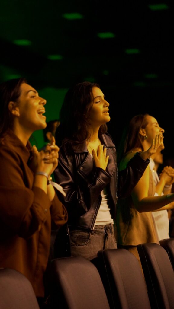 Três mulheres de pé, em adoração, entre as cadeiras do auditório