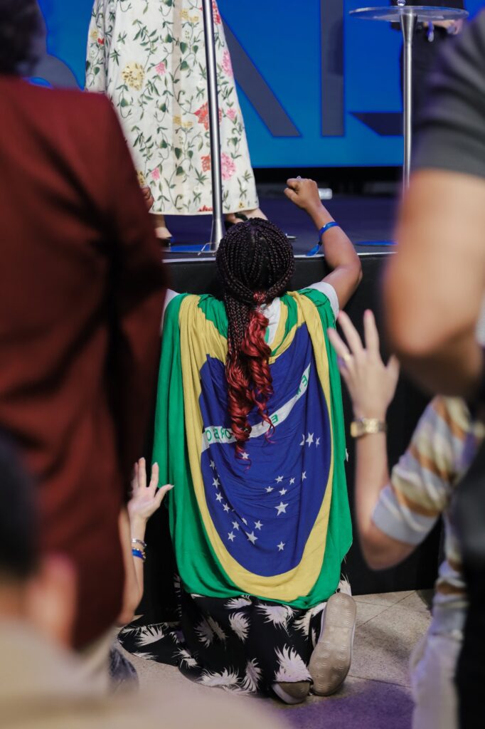 Mulher ajoelhada diante do palco, com a bandeira do Brasil nas costas, durante a oração na Conferência Global 2026