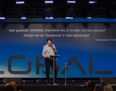 Tassos Lycurgo prega no palco da Conferência Global 2026, com Habacuque 1.2 projetado no telão