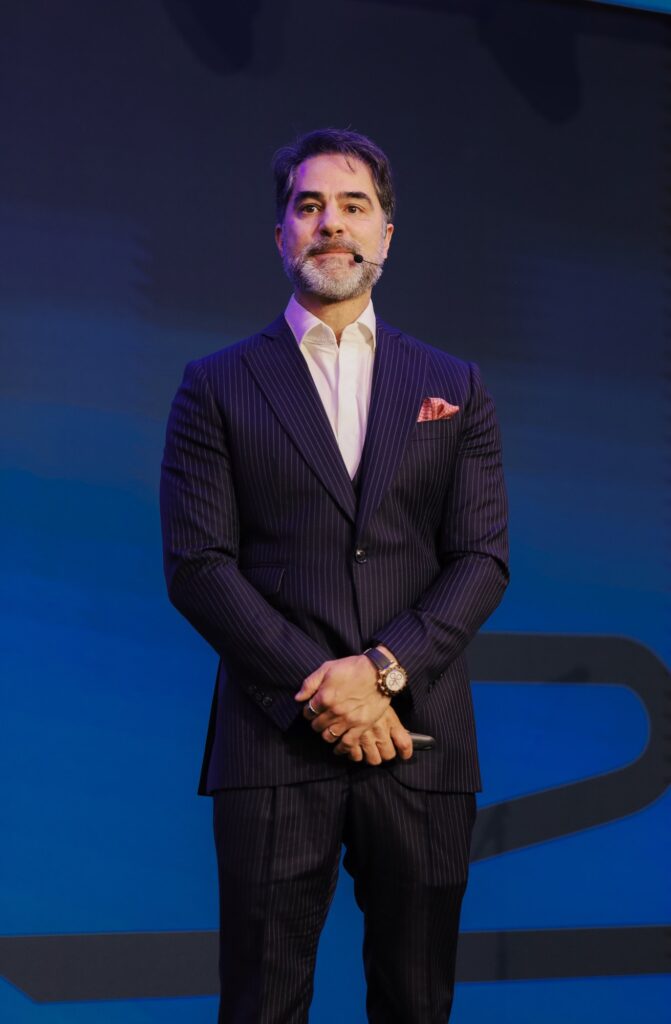 Victor Sorrentino de pé no palco, com as mãos juntas, diante do telão azul
