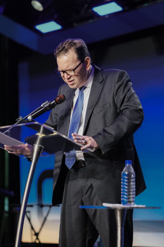 William Douglas lê suas anotações no púlpito, diante do telão azul da Conferência Global 2026